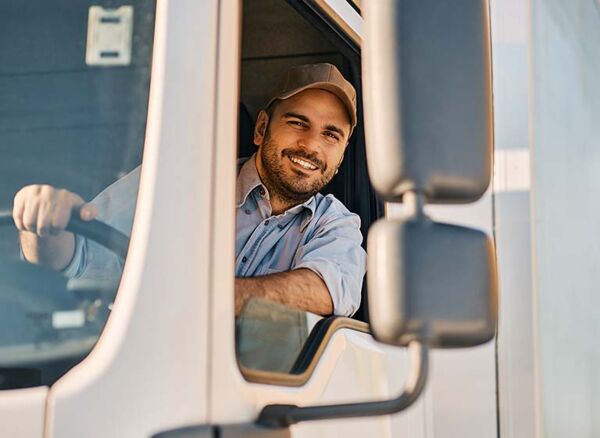 Mann hinter dem Lenkrad eines LKW lehnt sich zum Fenster raus und lächelt in die Kamera.